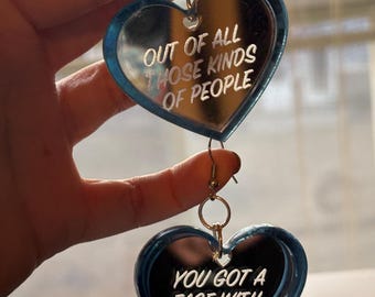 May include: A pair of blue heart-shaped earrings with silver hooks. The top earring has the text "Out of all those kinds of people" and the bottom earring has the text "You got a face with a view".