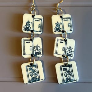 Op de afbeelding: Een paar opvallende oorbellen met vierkante, crèmekleurige hangers. Elke hanger heeft een cartoonillustratie van een persoon bij een deur. De deuren zijn in donkerblauw gelabeld met "WOMEN", "MEN" en "WHAT EVER". De oorbellen hebben zilverkleurige haken.