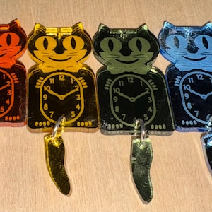 May include: Six cat-shaped acrylic charms in red, orange, yellow, green, blue, and purple. Each charm features a smiling cat face, a clock face on the body, and a separate dangling tail piece attached by a silver metal jump ring.