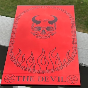 Puede incluir: Una placa rectangular roja con un grabado negro de un cráneo con cuernos centrado dentro de un borde de cadena. Debajo del cráneo hay llamas, y en la parte inferior, el texto THE DEVIL está flanqueado por dos símbolos de pentagrama.