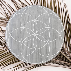 May include: A gray wooden round tray with a white geometric design, known as the Flower of Life, on top of a dried palm leaf.