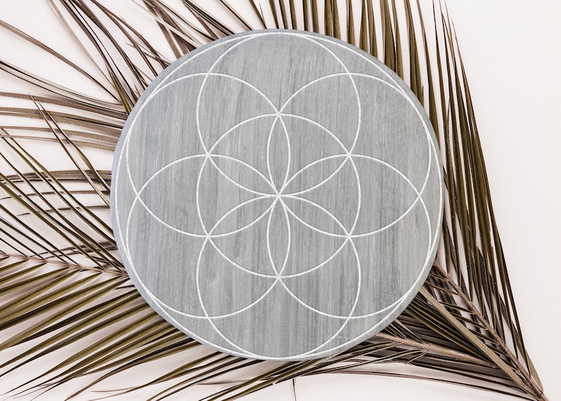 May include: A gray wooden round tray with a white geometric design, known as the Flower of Life, on top of a dried palm leaf.