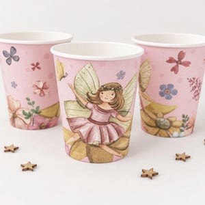May include: Three light pink paper cups decorated with watercolour illustrations of a fairy with wings, various flowers, and butterflies. Small wooden star-shaped confetti pieces are scattered around the base of the cups on a plain surface.