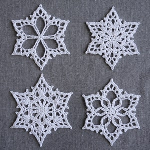 Peut inclure: Quatre flocons de neige en crochet blanc sur un fond de tissu gris. Les flocons de neige sont tous de designs diff&eacute;rents, mais ils sont tous faits avec le m&ecirc;me fil blanc.