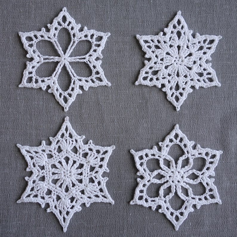 Peut inclure: Quatre flocons de neige en crochet blanc sur un fond de tissu gris. Les flocons de neige sont tous de designs diff&eacute;rents, mais ils sont tous faits avec le m&ecirc;me fil blanc.