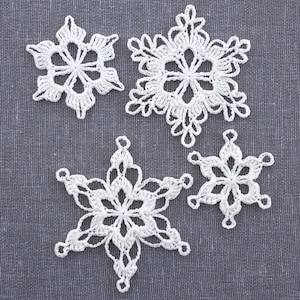 May include: Four white crocheted snowflakes on a gray fabric background. The snowflakes are delicate and intricate, with a six-pointed star shape.