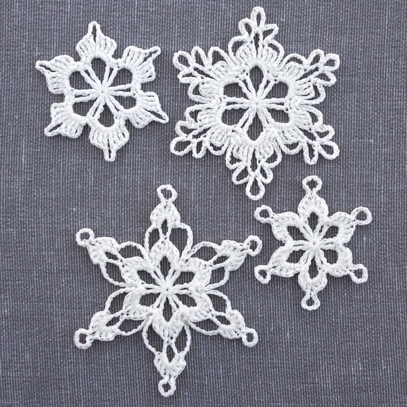 May include: Four white crocheted snowflakes on a gray fabric background. The snowflakes are delicate and intricate, with a six-pointed star shape.