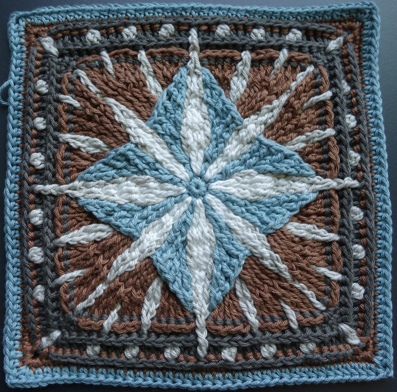 May include: A crocheted square with a starburst design in shades of brown, blue, and white. The center of the starburst is a white circle surrounded by blue and white stripes. The outer edges of the square are a dark gray border with white dots.