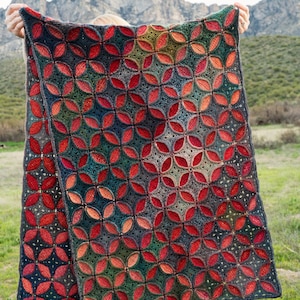 以下が含まれることがあります： 赤、緑、青の色合いで幾何学模様が施された、かぎ針編み毛布。毛布は山脈の前で持ち上げられている。