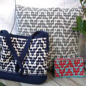 Puede incluir: Tres bolsas de crochet de diferentes tamaños, una bolsa de mano, una almohada y una pequeña bolsa. La bolsa de mano es azul marino y blanca, la almohada es gris y blanca, y la pequeña bolsa es roja y turquesa.