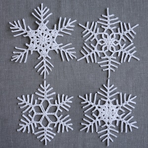 Peut inclure: Quatre flocons de neige en crochet blanc sur un fond de tissu gris. Les flocons de neige sont d&eacute;licats et complexes, avec une forme d'&eacute;toile &agrave; six branches.