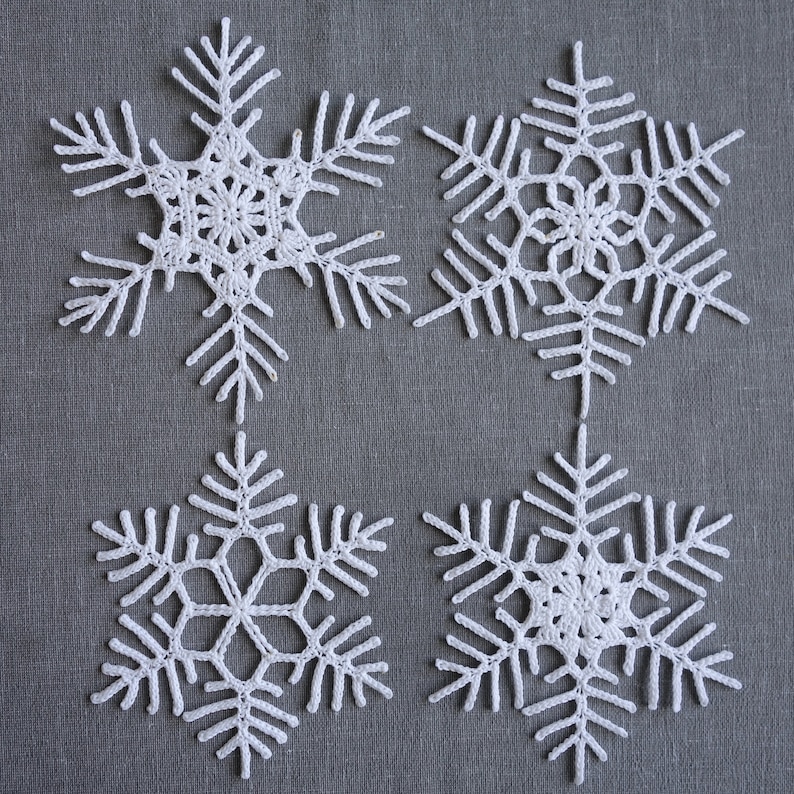 Peut inclure: Quatre flocons de neige en crochet blanc sur un fond de tissu gris. Les flocons de neige sont d&eacute;licats et complexes, avec une forme d'&eacute;toile &agrave; six branches.