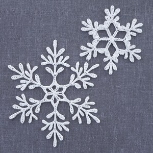 May include: Two white crocheted snowflakes on a gray fabric background.