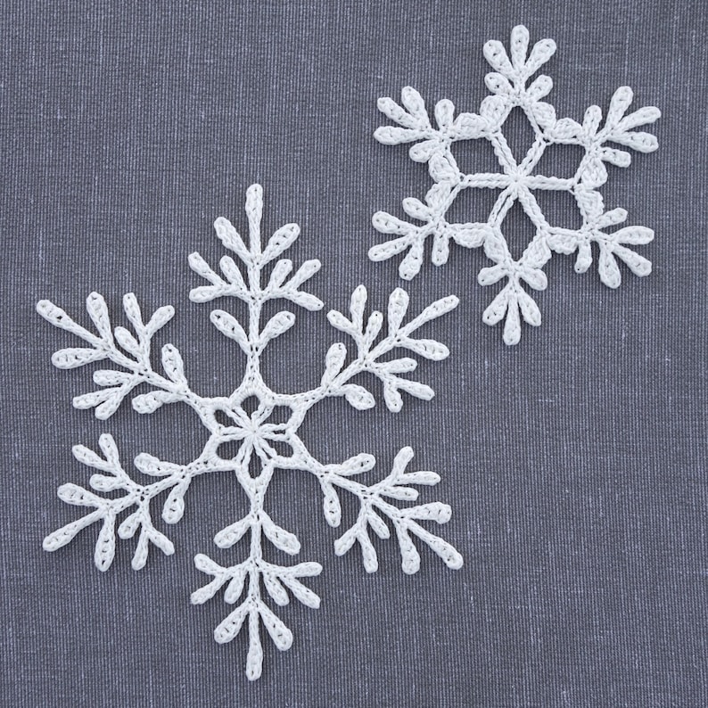 May include: Two white crocheted snowflakes on a gray fabric background.