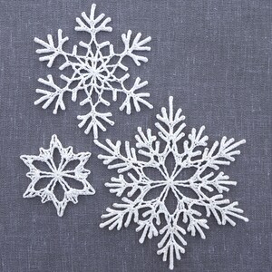 May include: Three white crocheted snowflakes of different sizes on a gray fabric background.