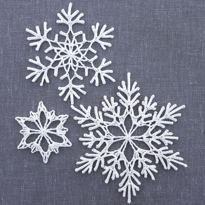 May include: Three white crocheted snowflakes of different sizes on a gray fabric background.