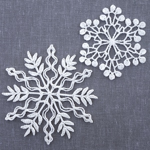May include: Two white crocheted snowflakes on a gray fabric background. The snowflakes are delicate and intricate, with a six-pointed star shape.