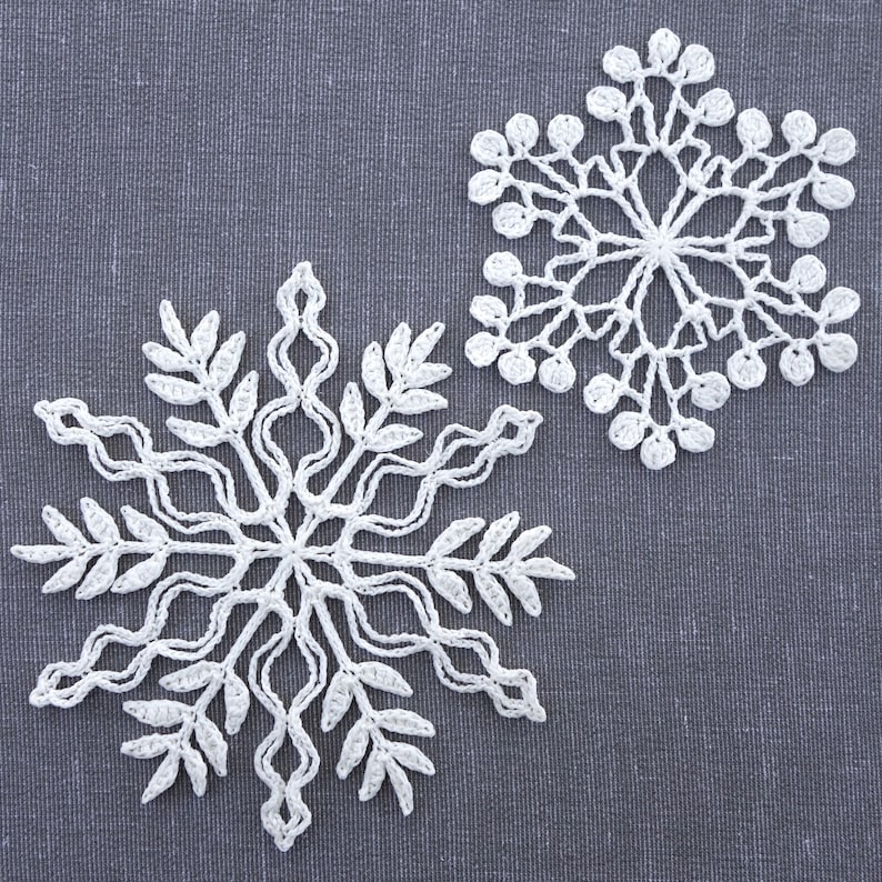 May include: Two white crocheted snowflakes on a gray fabric background. The snowflakes are delicate and intricate, with a six-pointed star shape.