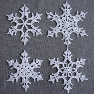 Peut inclure: Quatre flocons de neige en crochet blanc sur un fond de tissu gris. Les flocons de neige sont d&eacute;licats et complexes, avec une forme d'&eacute;toile &agrave; six branches.