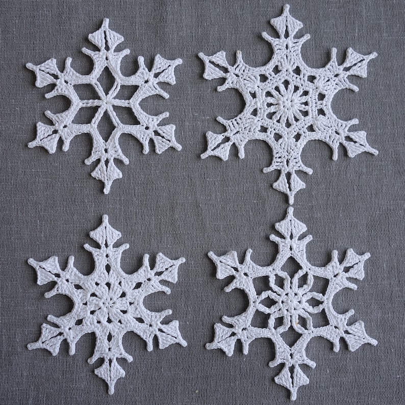 Peut inclure: Quatre flocons de neige en crochet blanc sur un fond de tissu gris. Les flocons de neige sont d&eacute;licats et complexes, avec une forme d'&eacute;toile &agrave; six branches.