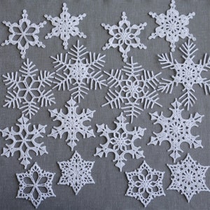 Peut inclure: Une collection de 15 flocons de neige en crochet blanc de diff&eacute;rentes tailles et motifs. Les flocons de neige sont dispos&eacute;s sur un fond de tissu gris.