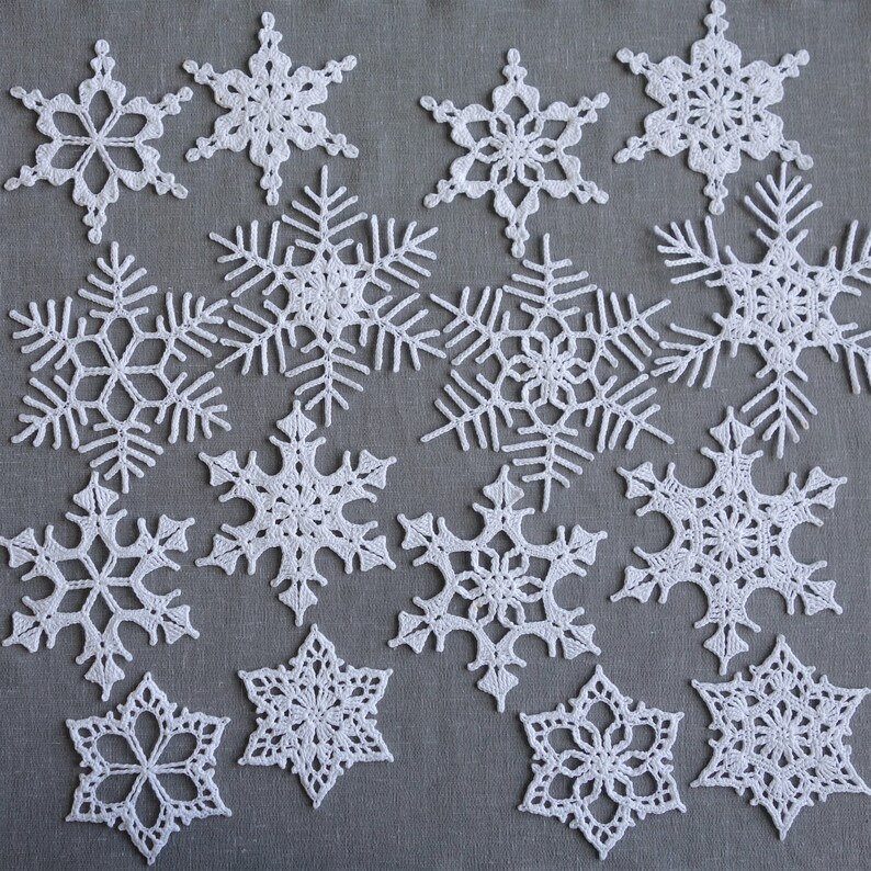Peut inclure: Une collection de 15 flocons de neige en crochet blanc de diff&eacute;rentes tailles et motifs. Les flocons de neige sont dispos&eacute;s sur un fond de tissu gris.
