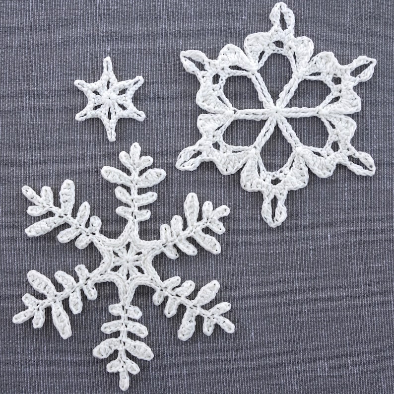May include: Three white crocheted snowflakes of varying sizes. The largest snowflake is in the center of the image, with a smaller snowflake to the left and a tiny snowflake to the upper right.