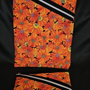 May include: Two zippered pouches with a vibrant orange floral pattern. The design features orange flowers with yellow centers on a black background. Each pouch has a black and silver diagonal stripe and a silver zipper.