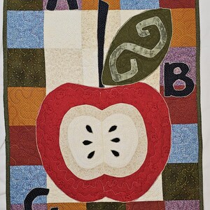 May include: A patchwork wall hanging with a large red apple design, cream-coloured centre, and black seeds. The letters A, B, and C are included, along with a green leaf. The background is a patchwork of various colours and patterns.