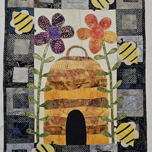 May include: A quilted wall hanging featuring a beehive, flowers, and bees. The beehive is brown and yellow with a black entrance. The flowers are purple, orange, and red. The bees are yellow and black with black stripes.