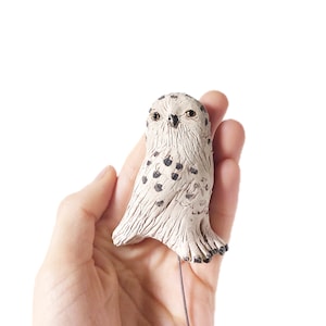 May include: A white ceramic owl figurine with black spots, perched on a thin wire. The owl has a detailed face with black eyes and a small beak.