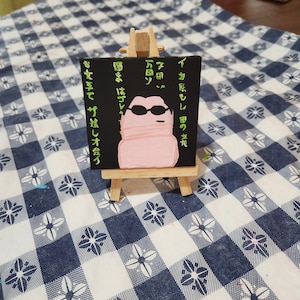 May include: A small black canvas painting on an easel with a pink cartoon character wearing sunglasses. The text on the canvas reads "イカもし田の文前 7日 万図ソ 図まはプレ 女手で ザ拭しオ会づ".