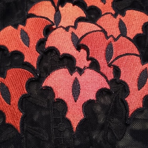 May include: A close-up of several red felt patches shaped like bats with black embroidered eyes. The patches are arranged in a layered pattern on a black lace background.