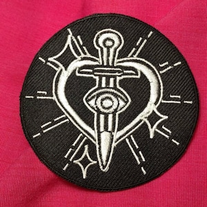 K&ouml;nnte beinhalten: Ein gestickter Patch in Schwarz und Wei&szlig; mit einem Dolch, der ein Herz durchbohrt. Das Herz hat ein Auge in der Mitte. Der Patch ist von einem Kreis aus wei&szlig;en N&auml;hten umgeben.