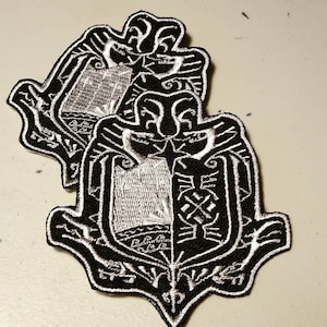 May include: Two black and white embroidered patches with a shield design. The shield is surrounded by a decorative border. The patches are layered on top of each other.