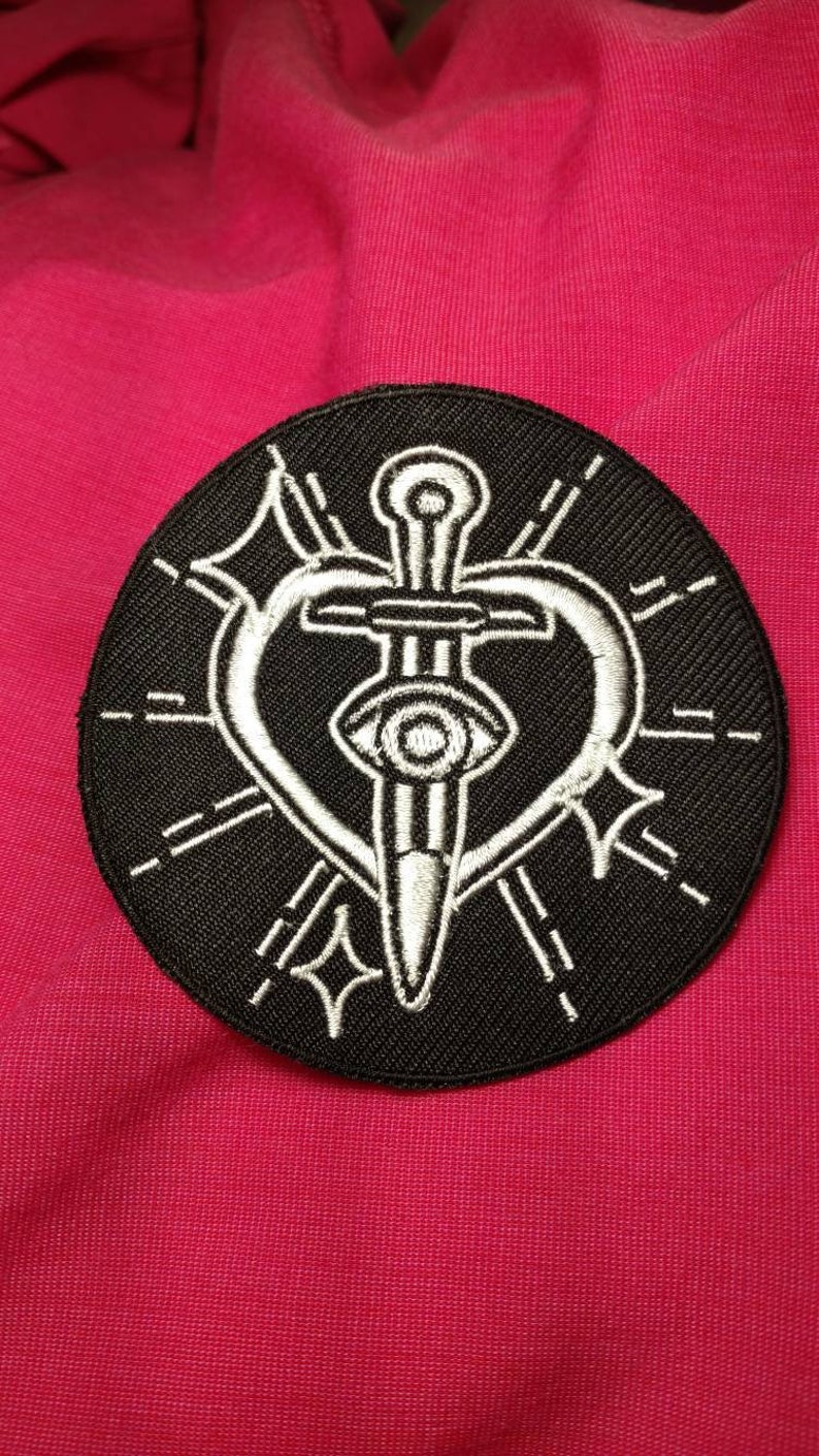 K&ouml;nnte beinhalten: Ein gestickter Patch in Schwarz und Wei&szlig; mit einem Dolch, der ein Herz durchbohrt. Das Herz hat ein Auge in der Mitte. Der Patch ist von einem Kreis aus wei&szlig;en N&auml;hten umgeben.