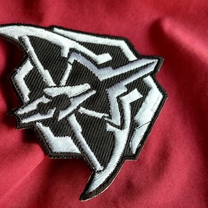 May include: A black and white embroidered patch with a geometric design. The patch is shaped like a shield and features a stylized star in the center.