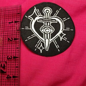 K&ouml;nnte beinhalten: Schwarz-wei&szlig; gestickter Patch mit einem Dolch, der ein Herz mit einem Auge in der Mitte durchbohrt. Das Herz ist von einem Kreis aus strahlenden Linien umgeben.