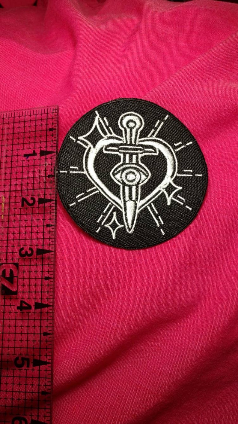 K&ouml;nnte beinhalten: Schwarz-wei&szlig; gestickter Patch mit einem Dolch, der ein Herz mit einem Auge in der Mitte durchbohrt. Das Herz ist von einem Kreis aus strahlenden Linien umgeben.