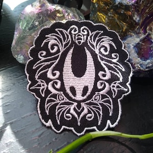 Puede incluir: Parche bordado en blanco y negro con un insecto estilizado con intrincados detalles florales. El insecto tiene una cabeza grande y redonda y un cuerpo largo y delgado. Los detalles florales están dispuestos alrededor del insecto en un patrón simétrico.