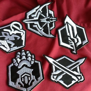 May include: Five black and white embroidered patches with different designs. The patches are hexagonal in shape and feature a variety of symbols, including swords, a claw, and a bow and arrow.