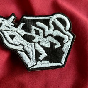 May include: White embroidered patch with a black background featuring a stylized scorpion design. The patch is shaped like a hexagon.