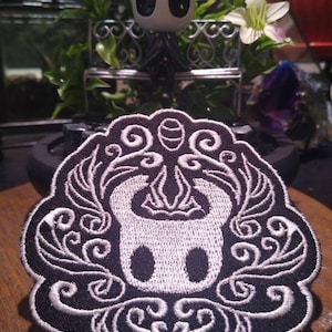 Könnte beinhalten: Ein gestickter Patch in Schwarzweiß mit einer stilisierten Silhouette des Ritters aus dem Videospiel Hollow Knight, umgeben von komplizierten floralen Designs.