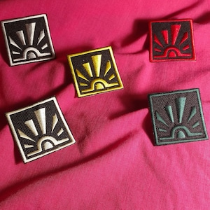 May include: Five embroidered patches with a sunrise design. The patches are black with different colored embroidery: white, yellow, red, green, and black.