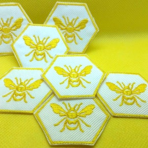 K&ouml;nnte beinhalten: Ein Set aus sieben wei&szlig;en, sechseckigen Patches mit gelben gestickten Bienen. Die Patches sind in einem Wabenmuster angeordnet.