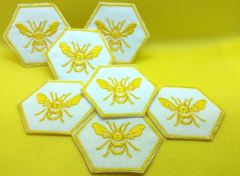 K&ouml;nnte beinhalten: Ein Set aus sieben wei&szlig;en, sechseckigen Patches mit gelben gestickten Bienen. Die Patches sind in einem Wabenmuster angeordnet.
