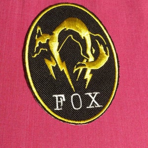 May include: A black and yellow embroidered patch featuring a stylised fox design and the word "FOX" in white lettering.