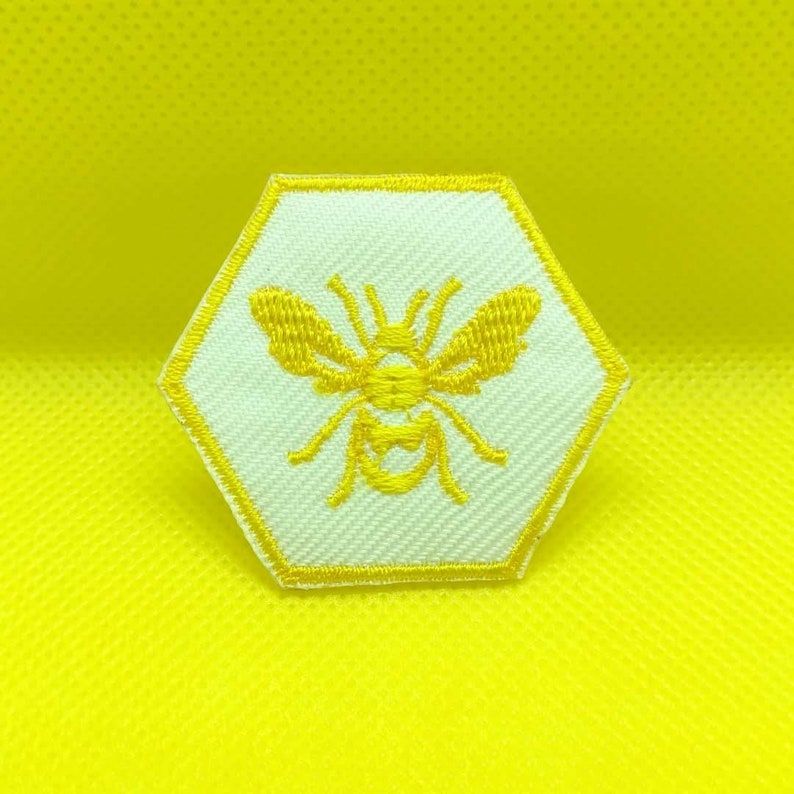 K&ouml;nnte beinhalten: Ein gelber gestickter Bienen-Patch in Form eines Sechsecks auf wei&szlig;em Hintergrund.