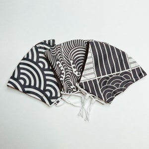 May include: Three black and white face masks with different geometric patterns. Each mask has a white adjustable string. The patterns include curved lines, waves, and vertical stripes. The masks are made of fabric.