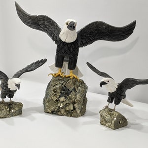 May include: Three carved bald eagle figurines perched on small, rough, gold-colored rock bases. The eagles are black with white heads and beaks.